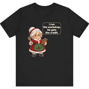 K&ouml;nnte beinhalten: Ein schwarzes T-Shirt mit einer Cartoon-Illustration einer &auml;lteren Frau in einem rot-wei&szlig;en Festtagsoutfit, die einen Holzl&ouml;ffel und ein Klemmbrett h&auml;lt. Eine Sprechblase enth&auml;lt den Text: I run the workshop. He gets the credit.