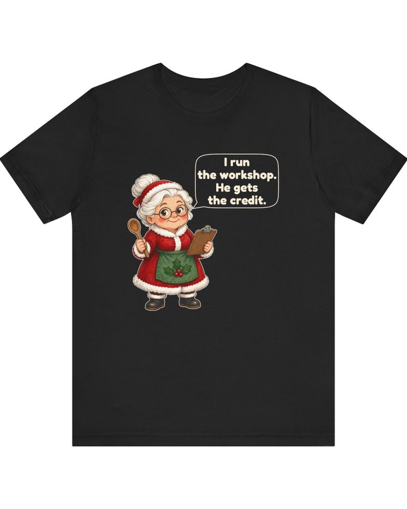 K&ouml;nnte beinhalten: Ein schwarzes T-Shirt mit einer Cartoon-Illustration einer &auml;lteren Frau in einem rot-wei&szlig;en Festtagsoutfit, die einen Holzl&ouml;ffel und ein Klemmbrett h&auml;lt. Eine Sprechblase enth&auml;lt den Text: I run the workshop. He gets the credit.