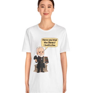 May include: A white t-shirt featuring a cartoon illustration of a bearded man in a suit holding a stack of books in front of a building. A speech bubble above him contains the text: Have you tried the library? I built a few.