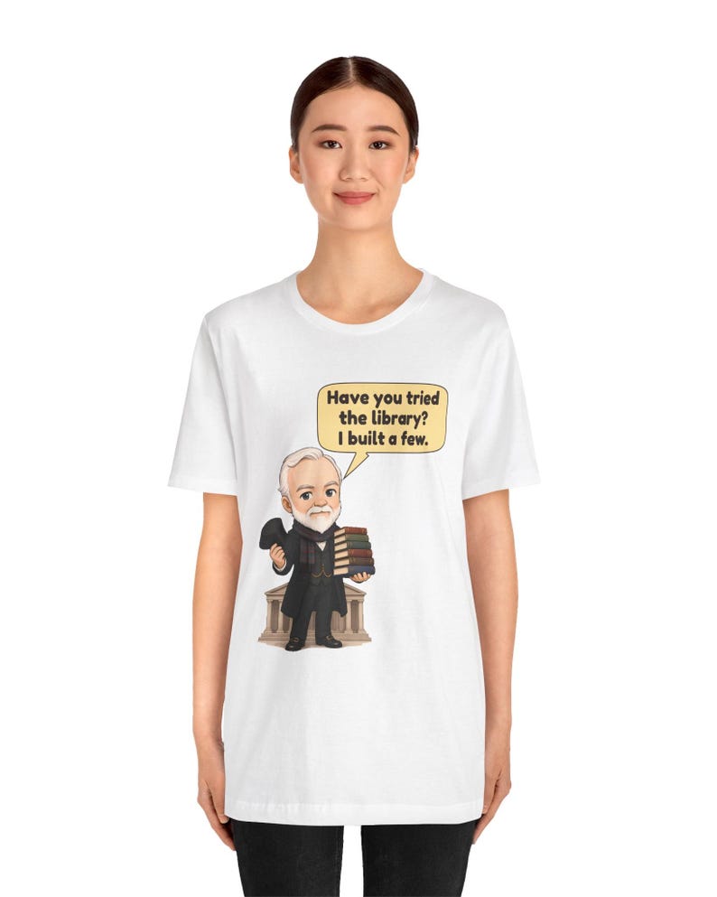 May include: A white t-shirt featuring a cartoon illustration of a bearded man in a suit holding a stack of books in front of a building. A speech bubble above him contains the text: Have you tried the library? I built a few.