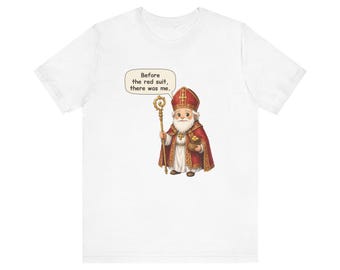 May include: A white t-shirt featuring a graphic of a Saint Nicholas figure in red and gold robes, holding a golden staff and a bag of coins. A speech bubble above the figure contains the text: Before the red suit, there was me.