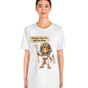 Może przedstawiać: Biały t-shirt z rysunkową ilustracją młodego faraona w niebiesko-złotym nakryciu głowy w paski, białej tunice i sandałach. Postać trzyma złote berło i bicz, z dymkiem z napisem, Mummy says I'm still the king.