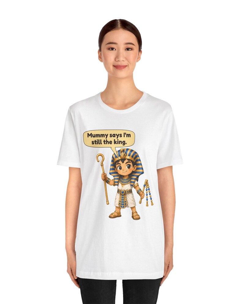 Może przedstawiać: Biały t-shirt z rysunkową ilustracją młodego faraona w niebiesko-złotym nakryciu głowy w paski, białej tunice i sandałach. Postać trzyma złote berło i bicz, z dymkiem z napisem, Mummy says I'm still the king.
