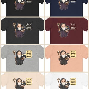 May include: A collection of eight t-shirts in various colors, each featuring a graphic of a bearded monk holding a staff and a book, with a raven perched on their shoulder. A speech bubble contains the text: Ora et labora. Mostly labora.