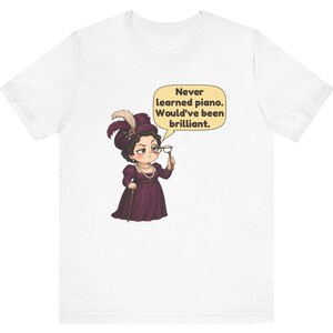 May include: A white t-shirt featuring a cartoon illustration of a person in a dark purple dress, hat with feathers, and pearls, holding a monocle. A speech bubble contains the text: Never learned piano. Would've been brilliant.