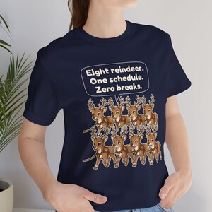 K&ouml;nnte beinhalten: Ein dunkelblaues Kurzarm-T-Shirt mit einer Grafik von acht braunen Rentieren in Geschirren. &Uuml;ber den Rentieren befindet sich eine wei&szlig;e Sprechblase mit dem Text: Eight reindeer. One schedule. Zero breaks. Das Shirt hat einen Rundhalsausschnitt.