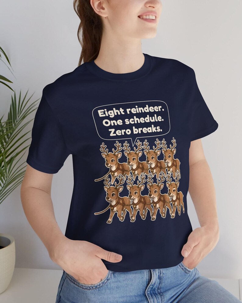 K&ouml;nnte beinhalten: Ein dunkelblaues Kurzarm-T-Shirt mit einer Grafik von acht braunen Rentieren in Geschirren. &Uuml;ber den Rentieren befindet sich eine wei&szlig;e Sprechblase mit dem Text: Eight reindeer. One schedule. Zero breaks. Das Shirt hat einen Rundhalsausschnitt.