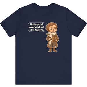 May include: A navy blue short-sleeve t-shirt featuring a cartoon illustration of a person in a brown coat and white scarf holding a quill and candle. A speech bubble next to the character contains the text: Underpaid, overworked, still festive.