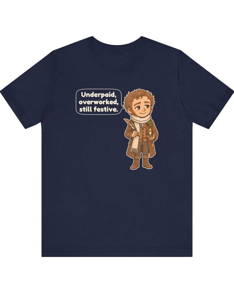 May include: A navy blue short-sleeve t-shirt featuring a cartoon illustration of a person in a brown coat and white scarf holding a quill and candle. A speech bubble next to the character contains the text: Underpaid, overworked, still festive.