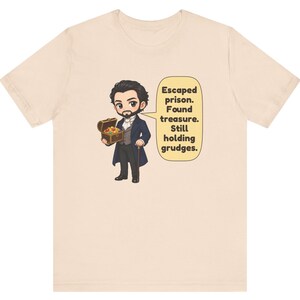 May include: A light beige t-shirt featuring a cartoon illustration of a bearded character in a dark blue coat holding a treasure chest. A speech bubble next to the character contains the text: Escaped prison. Found treasure. Still holding grudges.
