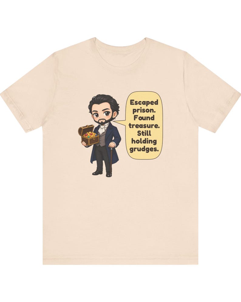 May include: A light beige t-shirt featuring a cartoon illustration of a bearded character in a dark blue coat holding a treasure chest. A speech bubble next to the character contains the text: Escaped prison. Found treasure. Still holding grudges.