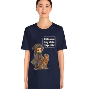 May include: A navy blue short-sleeve t-shirt featuring a graphic of a person in a tricorn hat and colonial-style clothing standing next to a small brown horse. A speech bubble above them contains the text: Delaware: tiny state. Huge ride.