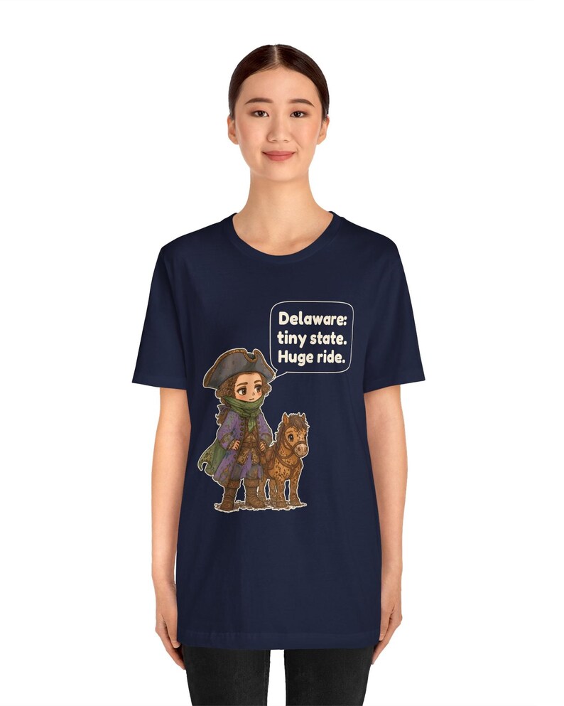May include: A navy blue short-sleeve t-shirt featuring a graphic of a person in a tricorn hat and colonial-style clothing standing next to a small brown horse. A speech bubble above them contains the text: Delaware: tiny state. Huge ride.