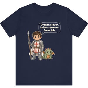 K&ouml;nnte beinhalten: Ein marineblaues T-Shirt mit einer Cartoon-Illustration eines Ritters in silberner R&uuml;stung, der einen Speer h&auml;lt und neben einem kleinen gr&uuml;nen Drachen steht. Eine Sprechblase enth&auml;lt den Text: Dragon slayer. Spider remover. Same job.