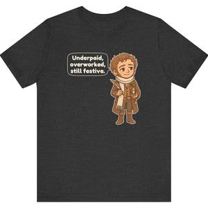 May include: A dark heather gray t-shirt featuring a cartoon illustration of a person in a brown coat and white scarf holding a quill and candle. A speech bubble contains the text: Underpaid, overworked, still festive.