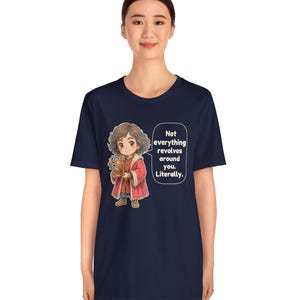 May include: A navy blue t-shirt featuring a cartoon illustration of a person with curly brown hair wearing a red coat and holding a golden globe. A speech bubble next to the character contains the text: Not everything revolves around you. Literally.