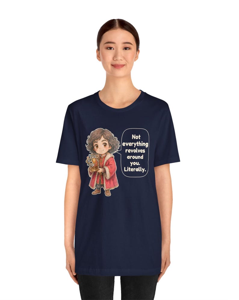 May include: A navy blue t-shirt featuring a cartoon illustration of a person with curly brown hair wearing a red coat and holding a golden globe. A speech bubble next to the character contains the text: Not everything revolves around you. Literally.