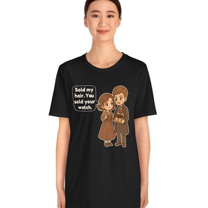 May include: A black short-sleeve t-shirt featuring a cartoon illustration of two figures. One figure holds a box, and a speech bubble contains the text: Sold my hair. You sold your watch.