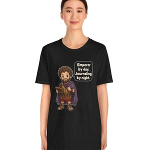 K&ouml;nnte beinhalten: Ein schwarzes T-Shirt mit einer Cartoon-Illustration eines b&auml;rtigen Mannes in r&ouml;mischer R&uuml;stung und violettem Umhang, der ein Tagebuch und einen Stift h&auml;lt. Eine Sprechblase neben der Figur enth&auml;lt den Text: Emperor by day. Journaling by night.