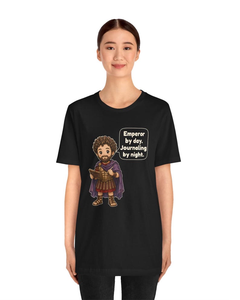 K&ouml;nnte beinhalten: Ein schwarzes T-Shirt mit einer Cartoon-Illustration eines b&auml;rtigen Mannes in r&ouml;mischer R&uuml;stung und violettem Umhang, der ein Tagebuch und einen Stift h&auml;lt. Eine Sprechblase neben der Figur enth&auml;lt den Text: Emperor by day. Journaling by night.
