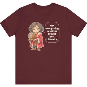 May include: A maroon short-sleeve t-shirt featuring a cartoon illustration of a person with curly hair wearing a red coat and holding a golden armillary sphere. A speech bubble contains the text: Not everything revolves around you. Literally.