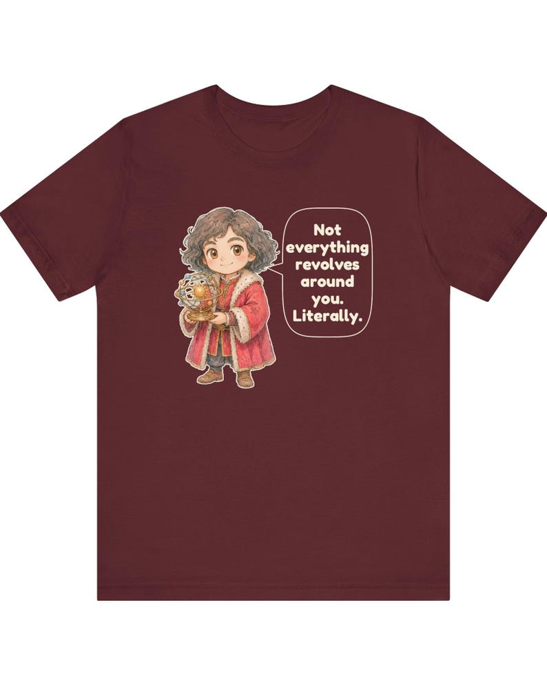 May include: A maroon short-sleeve t-shirt featuring a cartoon illustration of a person with curly hair wearing a red coat and holding a golden armillary sphere. A speech bubble contains the text: Not everything revolves around you. Literally.