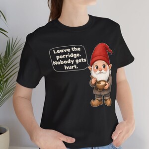 May include: A black t-shirt featuring a graphic of a gnome wearing a red hat and brown clothing, holding a bowl of porridge with a spoon. A white speech bubble next to the gnome contains the text: Leave the porridge. Nobody gets hurt.