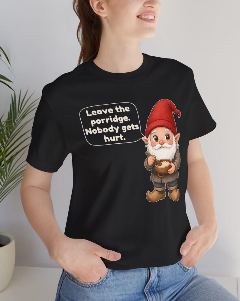 May include: A black t-shirt featuring a graphic of a gnome wearing a red hat and brown clothing, holding a bowl of porridge with a spoon. A white speech bubble next to the gnome contains the text: Leave the porridge. Nobody gets hurt.