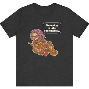 Puede incluir: Una camiseta gris oscuro de manga corta con el gr&aacute;fico de una persona mayor con pa&ntilde;uelo y gafas montada en una escoba. El gr&aacute;fico incluye un bocadillo con el texto: "Sweeping in late. Fashionably."