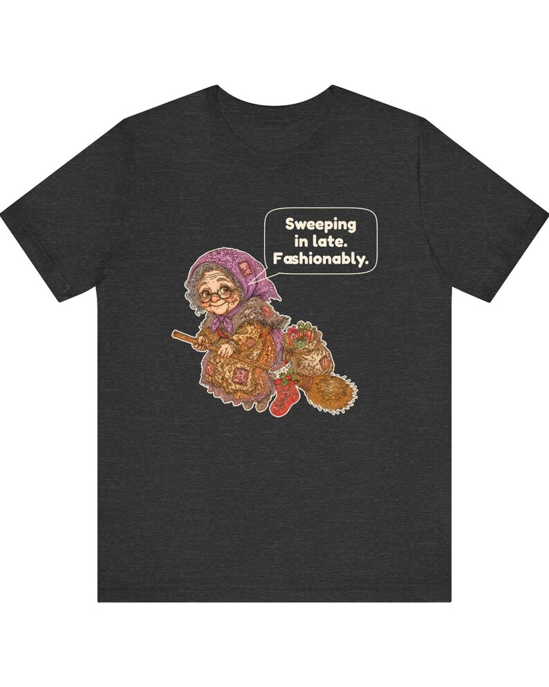 Puede incluir: Una camiseta gris oscuro de manga corta con el gr&aacute;fico de una persona mayor con pa&ntilde;uelo y gafas montada en una escoba. El gr&aacute;fico incluye un bocadillo con el texto: "Sweeping in late. Fashionably."