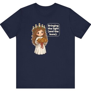 May include: A navy blue t-shirt featuring a graphic of a character wearing a white robe and a crown of candles with greenery, holding a tray of buns. A speech bubble next to the character contains the text: Bringing the light (and the buns).