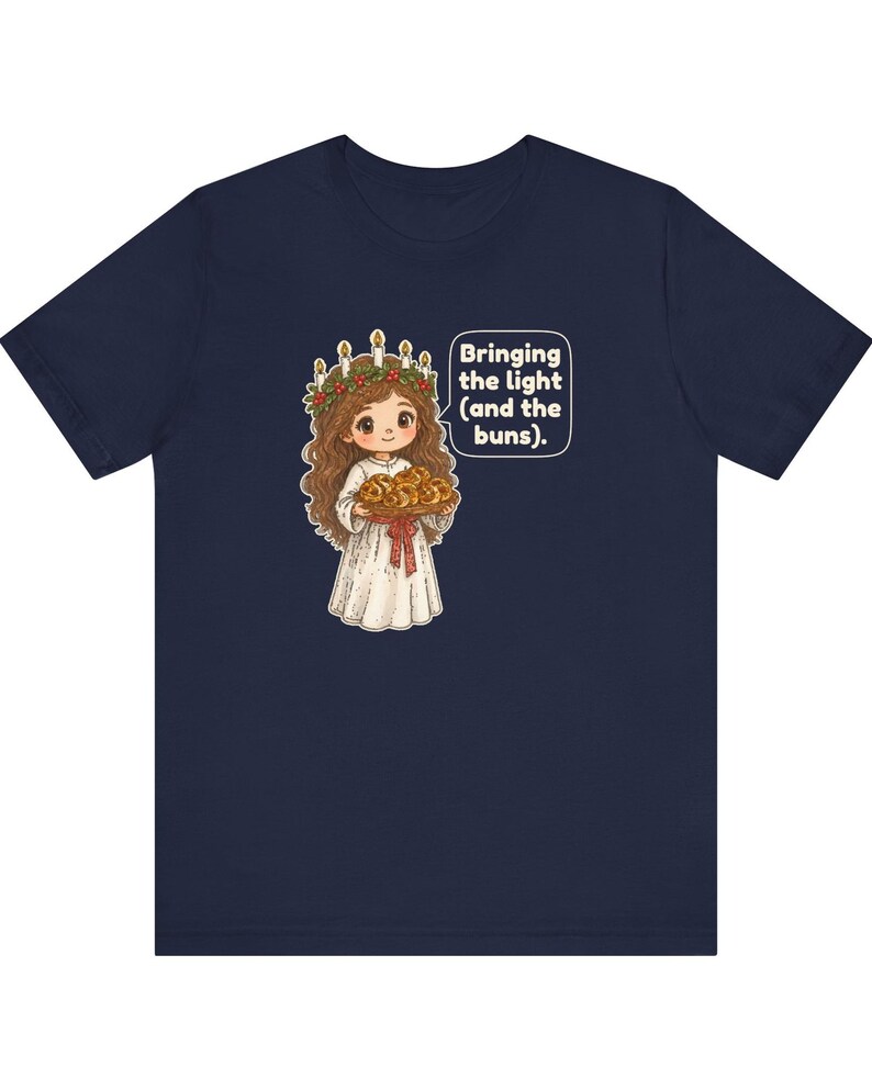 May include: A navy blue t-shirt featuring a graphic of a character wearing a white robe and a crown of candles with greenery, holding a tray of buns. A speech bubble next to the character contains the text: Bringing the light (and the buns).