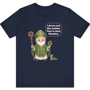 Puede incluir: Una camiseta azul marino con una ilustraci&oacute;n de dibujos animados de una figura con vestiduras religiosas verdes que sostiene un bast&oacute;n de madera y un tr&eacute;bol. Hay una peque&ntilde;a serpiente cerca y un globo de texto dice: I drove out the snakes. You're next, Monday.
