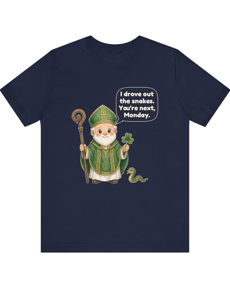 Puede incluir: Una camiseta azul marino con una ilustraci&oacute;n de dibujos animados de una figura con vestiduras religiosas verdes que sostiene un bast&oacute;n de madera y un tr&eacute;bol. Hay una peque&ntilde;a serpiente cerca y un globo de texto dice: I drove out the snakes. You're next, Monday.