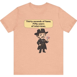 Pu&ograve; includere: Una t-shirt color pesca con un'illustrazione a cartoni animati di una figura con baffi, cappello e cappotto neri che tiene in mano delle carte da gioco. Un fumetto sopra la figura contiene il testo: Thirty seconds of fame. Fifty years of interviews.