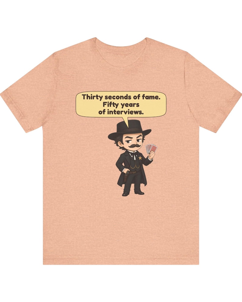 Pu&ograve; includere: Una t-shirt color pesca con un'illustrazione a cartoni animati di una figura con baffi, cappello e cappotto neri che tiene in mano delle carte da gioco. Un fumetto sopra la figura contiene il testo: Thirty seconds of fame. Fifty years of interviews.