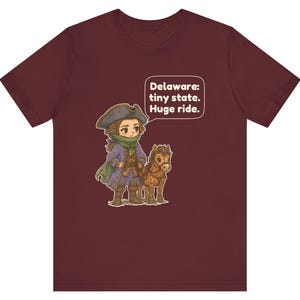 May include: A maroon short-sleeve t-shirt featuring a cartoon illustration of a person in a tricorn hat and colonial-style clothing standing next to a small brown horse. A speech bubble contains the text: Delaware: tiny state. Huge ride.