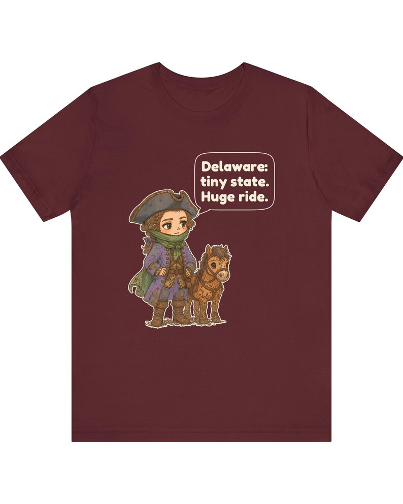 May include: A maroon short-sleeve t-shirt featuring a cartoon illustration of a person in a tricorn hat and colonial-style clothing standing next to a small brown horse. A speech bubble contains the text: Delaware: tiny state. Huge ride.