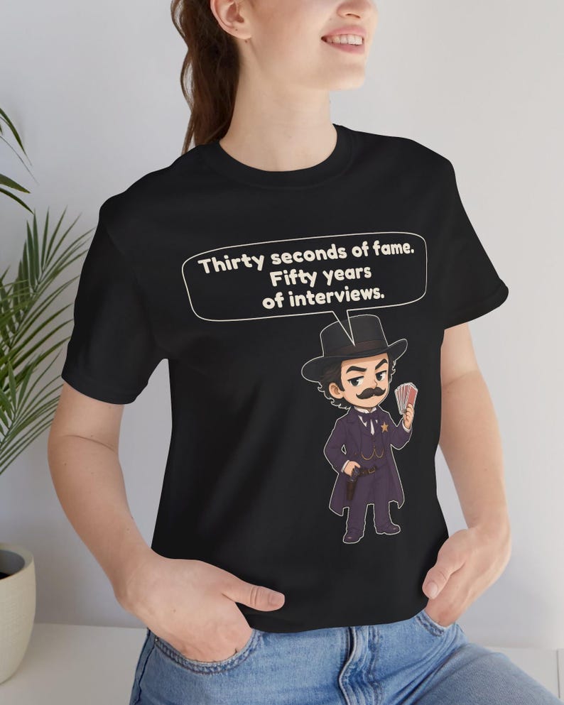 Pu&ograve; includere: Una t-shirt nera con un'illustrazione a cartone animato di un uomo in abito scuro, cappello e baffi che tiene in mano un mazzo di carte. Un fumetto sopra il personaggio contiene il testo: Thirty seconds of fame. Fifty years of interviews.