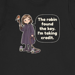 May include: A black t-shirt featuring a graphic of a person in a dark purple coat holding a large key with a robin perched on it. The person holds a rope with red and blue striped ends. A speech bubble contains the text: The robin found the key. I'm taking credit.