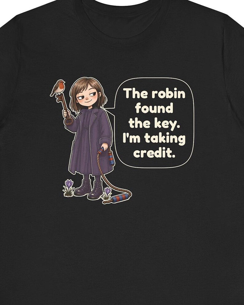 May include: A black t-shirt featuring a graphic of a person in a dark purple coat holding a large key with a robin perched on it. The person holds a rope with red and blue striped ends. A speech bubble contains the text: The robin found the key. I'm taking credit.
