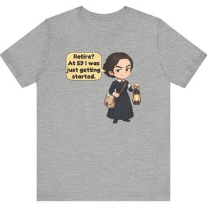 May include: A heather gray t-shirt featuring a cartoon illustration of a person in a dark dress carrying a satchel and a lit lantern. A speech bubble next to the character contains the text: Retire? At 59 I was just getting started.