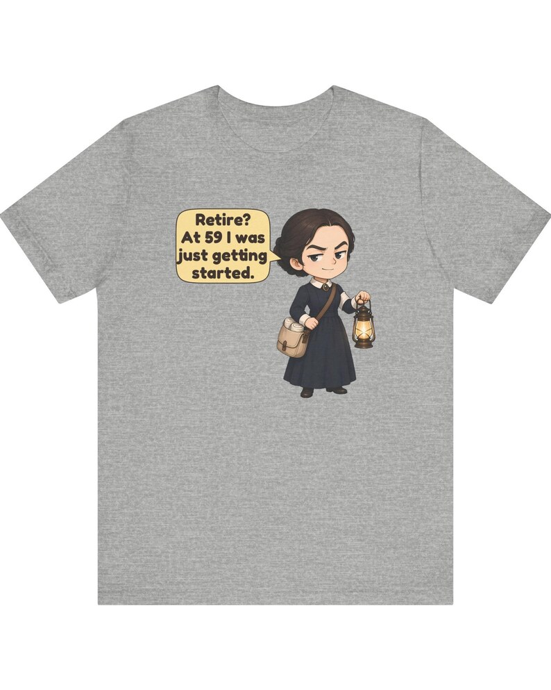 May include: A heather gray t-shirt featuring a cartoon illustration of a person in a dark dress carrying a satchel and a lit lantern. A speech bubble next to the character contains the text: Retire? At 59 I was just getting started.