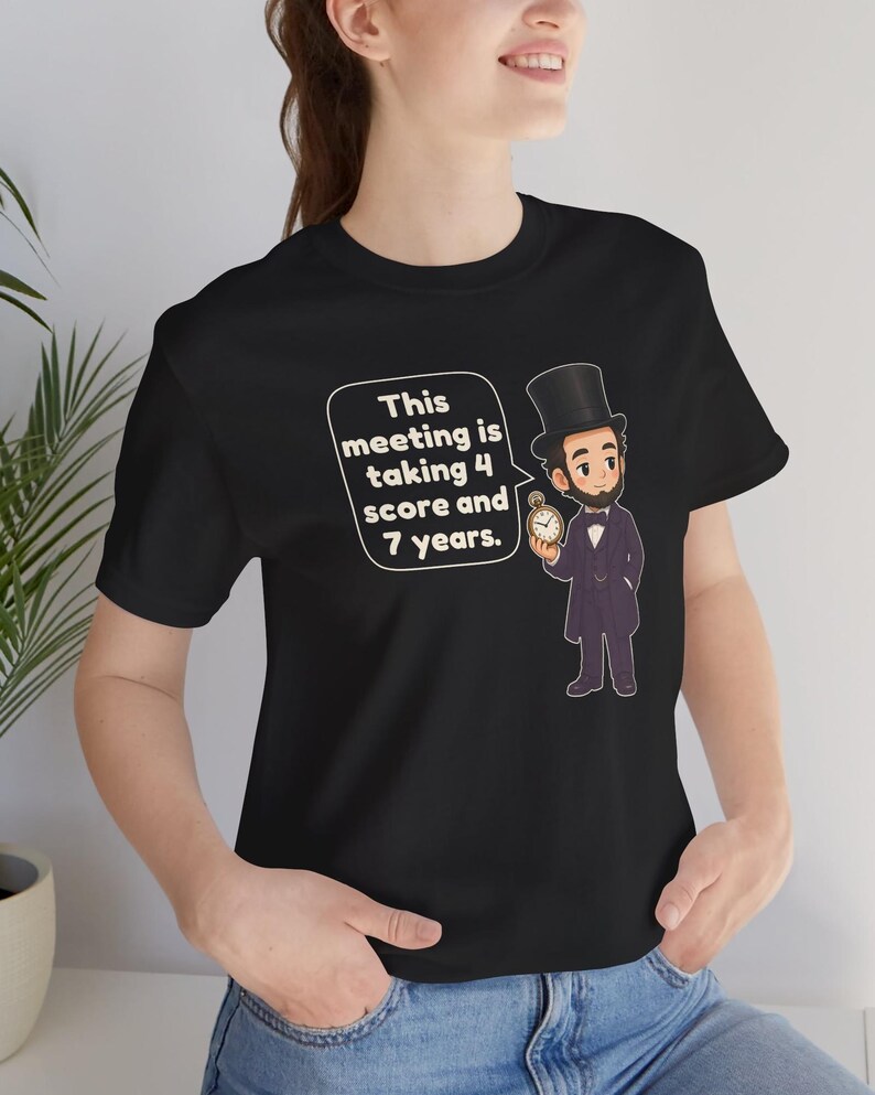 May include: A black short-sleeved t-shirt featuring a cartoon illustration of Abraham Lincoln wearing a top hat and holding a pocket watch. A speech bubble next to the figure contains the text: This meeting is taking 4 score and 7 years.