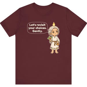 May include: A maroon t-shirt featuring a cartoon character with a candle flame on their head, holding a sprig of holly and a small gold pot. A speech bubble contains the text: Let's revisit your choices. Gently. The character wears a white tunic.