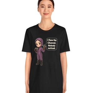 Puede incluir: Una camiseta negra con una ilustraci&oacute;n de estilo dibujo animado de una persona con un traje de vuelo morado y gafas. Un globo de texto junto al personaje contiene el texto: I flew the Channel. Nobody noticed. El dise&ntilde;o est&aacute; centrado.