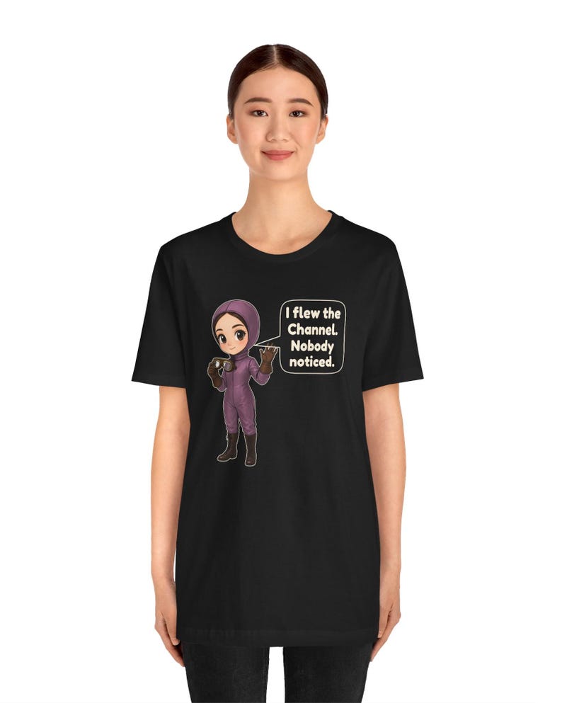 Puede incluir: Una camiseta negra con una ilustraci&oacute;n de estilo dibujo animado de una persona con un traje de vuelo morado y gafas. Un globo de texto junto al personaje contiene el texto: I flew the Channel. Nobody noticed. El dise&ntilde;o est&aacute; centrado.
