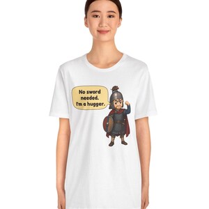May include: A white t-shirt featuring a cartoon illustration of a warrior in armor with a helmet, cape, and shield. A speech bubble next to the character contains the text: No sword needed. I'm a hugger. The warrior has a small animal on their helmet.