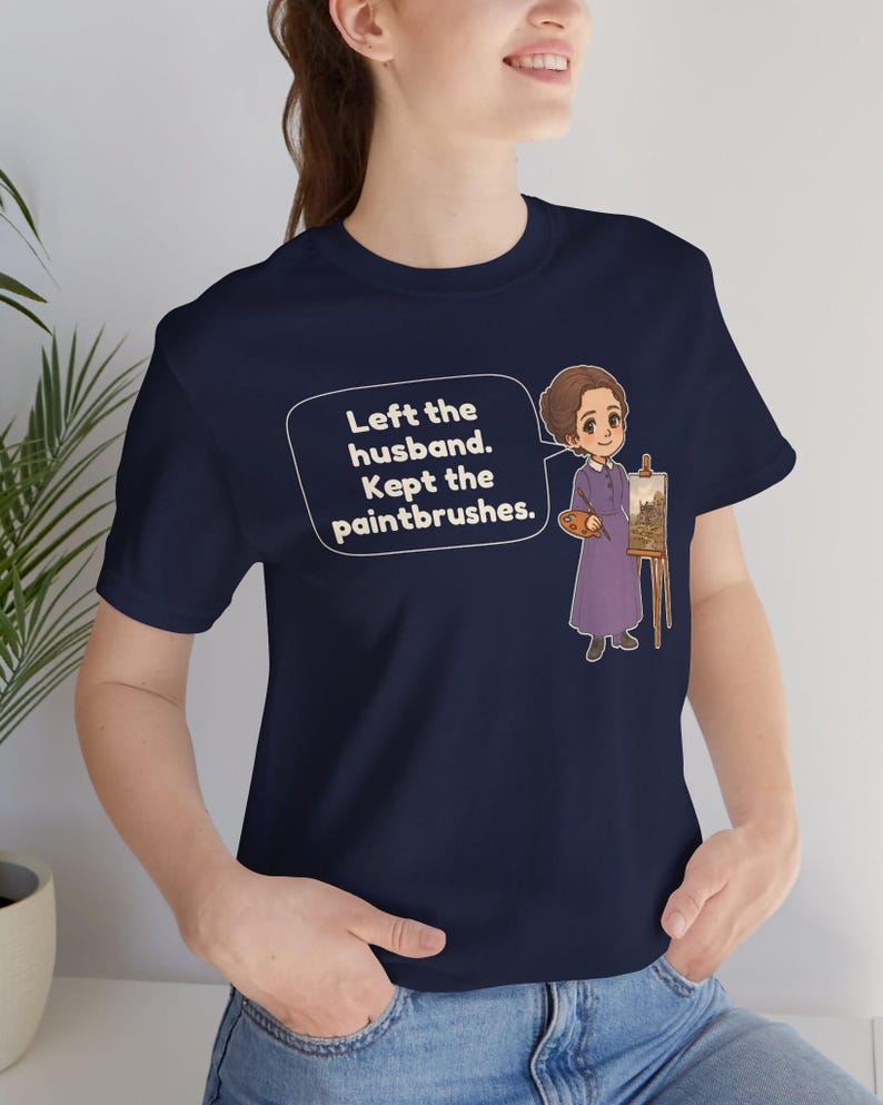 May include: A navy blue t-shirt featuring a cartoon illustration of a person in a purple dress standing by an easel with a paint palette. A speech bubble contains the text: Left the husband. Kept the paintbrushes.