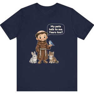 May include: A navy blue t-shirt featuring a cartoon illustration of a monk in a brown robe with a white rope belt. The monk is surrounded by a gray rabbit, a brown wolf, and a blue bird. A speech bubble contains the text: My pets talk to me. Yours too?